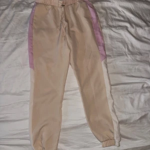 Beige mjukisbyxor med lila och vita detaljer från Amisu - Säljer ett par beige mjukisbyxor från Amisu med lila och vita paneler längs sidorna. Byxorna har resår i midjan med dragsko och resår vid bensluten. Perfekta för en avslappnad stil.