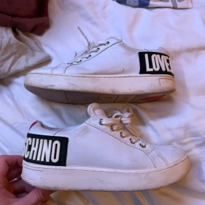 Vita sneakers från Moschino med textdetalj - Säljer ett par vita sneakers från Moschino med tydlig svartvit textdetalj på hälen där det står 'LOVE MOSCHINO' och ett rött hjärta. Skorna har vita snören och en platt sula. Perfekta för dig som gillar statement-plagg och streetstyle.