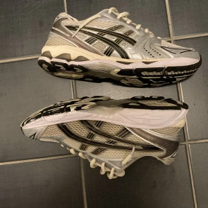 Asics gel kayano 14  - Säljer ett par silvriga Asics sneakers med svarta och vita detaljer. Skorna har snörning, mesh- och syntetmaterial samt en robust sula med svart och vit mönstring. Perfekta för träning eller vardagsstil. Storlek 42,5 men passar utmärkt på 41