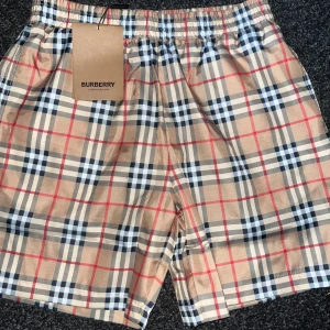 Rutig badshorts från Burberry - Snygga shorts från Burberry i klassiskt rutigt mönster med beige bas och inslag av svart, vitt och rött. Shortsen har resår i midjan och innerfoder i mesh. Perfekta för sommaren! Helt oanvända