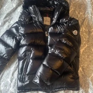 Moncler jacka  - Säljer en svart glansig dunjacka från Moncler med huva och dragkedja. Jackan har Moncler-logga på ärmen och är perfekt för kalla dagar. Klassisk pufferstil som håller värmen och ger en snygg look.