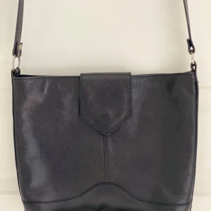 Svart axelväska i äkta skinn - Säljer en stilren svart axelväska i skinn med justerbart axelband. Väskan har ett lock med magnetknäppning och en praktisk ytterficka med dragkedja på baksidan. Perfekt för att bära med sig det viktigaste.