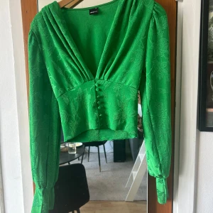 Grön blus med knappar från Gina Tricot - Säljer en grön blus från Gina Tricot med v-ringning, långa ärmar och fina klädda knappar framtill. Blusen har ett subtilt blommigt mönster och lätt glansigt tyg som ger en lyxig känsla. Perfekt för dig som vill sticka ut med färg!