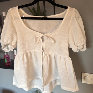 Vit blus med knyt från Gina Tricot - Säljer en söt vit blus från Gina Tricot med puffärmar och knyt framtill. Blusen har en lätt rynkad struktur, kort ärm med resår och volang nertill. Perfekt för vår och sommar!
