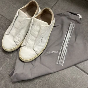 Vita sneakers från Axel Arigato - Säljer ett par vita sneakers från Axel Arigato i stilren design utan snörning. Skorna har rund tå och är tillverkade i skinn med diskret logga på sidan. Perfekta för en clean och minimalistisk look.