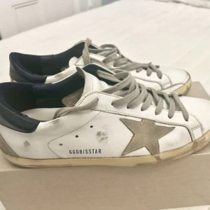 Vita sneakers med grå stjärna från Golden Goose - Säljer ett par vita sneakers från Golden Goose med grå stjärna på sidan, grå snörning och svarta detaljer vid hälen. Skorna har en klassisk låg modell och är perfekta för en avslappnad stil.