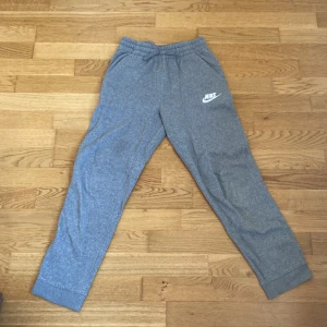 Grå mjukisbyxor från Nike - Grå mjukisbyxor från Nike med resår i midjan och muddar vid bensluten. Byxorna har två sidofickor och en vit Nike-logga tryckt på vänster lår. 