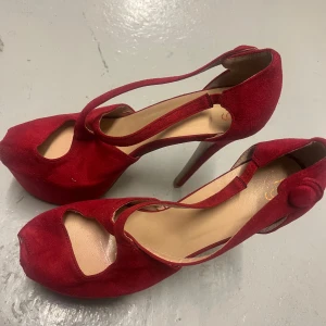 Röda höga pumps med öppen tå - Säljer ett par röda pumps med hög klack och öppen tå. Skorna har en snygg rem över foten och är tillverkade i mocka. Perfekta för dig som vill sticka ut med ett par eleganta klackar.