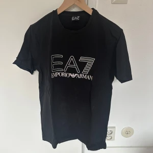 Svart t-shirt från Emporio Armani EA7 - Säljer en svart t-shirt från Emporio Armani EA7 med stort vitt logotryck på bröstet. Klassisk rund halsringning och korta ärmar. Perfekt för en stilren och sportig look.