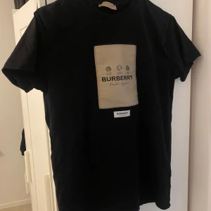 Svart t-shirt från Burberry med beige patch - Säljer en svart t-shirt från Burberry med en stor beige patch på bröstet där det står 'Burberry London, England'. T-shirten har klassisk passform och korta ärmar. Perfekt för dig som gillar stilrena märkeskläder.