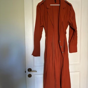 Rostfärgad trenchcoat från Zara - Säljer en rostfärgad långklänning från Zaras TRF Collection med knappar framtill och snöre i midjan. Mycket fina skick.
