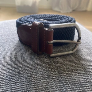 Blått flätat bälte med bruna skinn-detaljer - Snyggt blått flätat bälte med detaljer i brunt skinn och ett robust silverfärgat spänne. Perfekt accessoar för att lyfta din outfit med en klassisk touch.