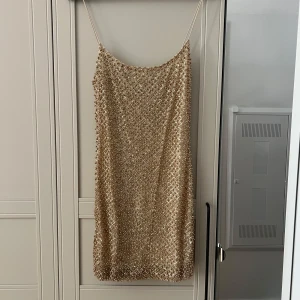 Glittrig guldig kortklänning från Oh Polly - Glittrig guldig klänning från Oh Polly med smala axelband. Klänningen är i väldigt fint skick och sitter som på modellen, sann i storlek. Den har tappat någon enstaka pärla här och där men inget som märks. Glittrar lite av sig. Upplever den inte så genomskinlig som på första bilden. 