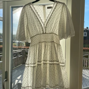 Vit kortklänning med spets från By Malina - Superfin vit kortklänning från By Malina med v-ringning, volangdetaljer och spets längs kanterna. Klänningen har puffiga kortärmar, smock i midjan och ett diskret prickigt mönster. Perfekt för sommarens alla tillfällen!