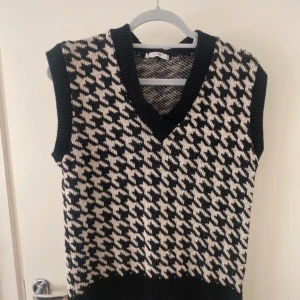 Svartvit stickad väst  - Snygg stickad väst i svart och vitt mönster och V-ringad och ribbade kanter vid hals, ärmhål och nederkant. Perfekt att styla över en skjorta eller t-shirt. 
