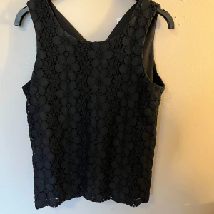 Svart spetsmönstrad linnetopp från Vero Moda - Säljer en svart ärmlös topp från Vero Moda med vackert blommigt spetsmönster. Toppen har rund halsringning och knäppning bak i nacken. Perfekt att styla till olika outfits!