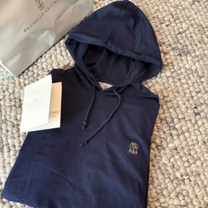 Brunello Cucinelli hoodie - Säljer en marinblå hoodie från Brunello Cucinelli i slim fit-modell. Tröjan är tillverkad i 100% bomull och har långa ärmar samt en liten broderad logga på bröstet. Perfekt för dig som gillar stilrena och exklusiva plagg.