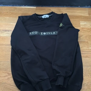 Svart sweatshirt från Stone Island - Svart sweatshirt från Stone Island med klassisk logotyp på bröstet och patch på ärmen. Tröjan har rund halsringning och ribbade muddar vid ärmslut och nederkant. Perfekt för en stilren och avslappnad look.