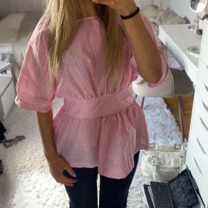 Rosa och vit randig blus med rosett i ryggen - INTRESSEKOLL! Säljer en söt rosa och vit randig blus med bred midjedel och knytrosett i ryggen. Blusen har korta ärmar och en öppen rygg, vilket ger en unik och trendig look. Perfekt för vår och sommar!  , man spänner i midjan bakifrån och passar därför olika strlk!!❤️