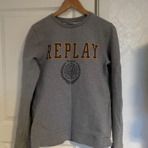 Grå sweatshirt från Replay - Säljer en grå sweatshirt från Replay med tryckt logga och emblem på bröstet. Perfekt för en stilren look. Storlek M säljer för 400kr men pris kan diskuteras vid snabb affär. Tröjan går inte att beställa längre och den är knappt använd.