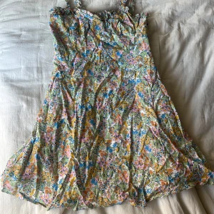 Blommig sommarklänning från Zara - Supersöt blommig klänning från Zara med smock upptill och volangiga axelband. Klänningen har en ljus bas med färgglada blommor i rosa, blått, gult och grönt. Perfekt för varma dagar och midsommarfirande.