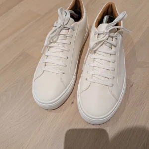 NYA Vita sneakers med snörning stl 42 - Stilrena vita sneakers från Zara med rund tå och platt sula. Skorna har klassisk snörning och är tillverkade i syntetmaterial med en ljusbrun insida. Perfekta för en clean och modern look.