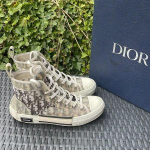 Dior B23  - Säljer ett par Dior B23 high top sneakers med ikoniskt monogrammönster i grått och vitt. Skorna har genomskinliga paneler, vita snören och en platt sula med Dior-logga på sidan. Perfekta för dig som vill sticka ut med lyxig streetstyle.