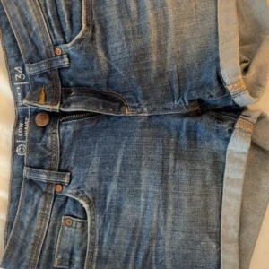 Blå jeansshorts low waist - Säljer ett par klassiska blå jeansshorts med låg midja och upprullade benslut. Shortsen har fem fickor och stängs med knapp och dragkedja. Perfekta för sommaren och enkla att matcha med olika toppar.