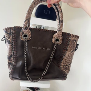 Brun handväska med ormmönster från Stella McCartney - Säljer en snygg brun handväska från Stella McCartney med ormmönstrade detaljer och kedjehandtag. Perfekt för att lyfta din outfit. priset kan diskuteras💗💗💗