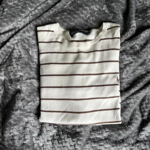 Vit randig tröja från Polo Ralph Lauren - Vit långärmad tröja från Polo Ralph Lauren med bruna horisontella ränder och broderad logga på bröstet. Klassisk rund halsringning och ribbade muddar. Perfekt för en stilren och avslappnad look. Bara att höra av sig för fler bilder eller funderingar.