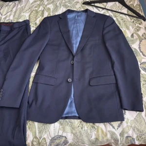 Mörkblå kostym med kavaj och byxor - Mörkblå kostym från Dressmann – stl 46 Regular / byxa 33/32 Slimfit (nyskick)  Säljer en klassisk och stilren tvådelad kostym från Dressmann, perfekt till student, bröllop, bal eller andra högtidliga tillfällen. Kostymen är i nyskick – endast använd en gång.  Märke: Dressmann  Färg: Mörkblå  Modell: Tvåknäppt kavaj med dubbla sprund och fickor fram  Storlek kavaj: 46 Regular  Storlek byxa: 33/32 Slimfit  Passform: Modern och välsittande  fluga ingår ej Skick: Nyskick – endast använd en 