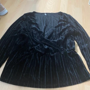 Svart omlottopp i sammet från ONLY - Säljer en svart omlottopp i mjuk sammet från ONLY. Toppen har lång ärm och v-ringning, perfekt för att lyfta din outfit med en lyxig känsla. Snyggt draperad framtill och passar till många olika stilar.