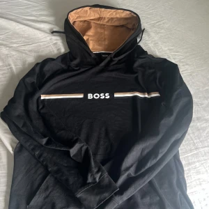 Svart hoodie från BOSS med beige detaljer - Säljer en svart hoodie från BOSS med beige och vita ränder över bröstet och texten 'BOSS' i vitt. Insidan av huvan är beige vilket ger en snygg kontrast. Perfekt för en avslappnad stil. Nypris för 1059kr. Kvitto finns!
