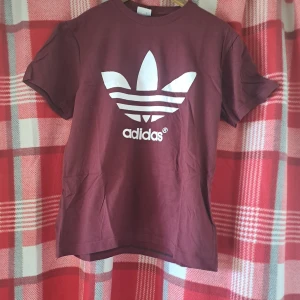 Vinröd t-shirt från Adidas - Säljer en vinröd t-shirt från Adidas med det klassiska vita Trefoil-loggan tryckt på bröstet. T-shirten har rund halsringning och korta ärmar. Perfekt för en sportig och avslappnad stil.