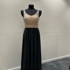 Svart långklänning med glittrig topp - Elegant långklänning med svart kjol och glittrig, ljusbeige topp dekorerad med stenar. Ärmlös modell med rundad ringning, perfekt för bal eller fest. Kjolen faller mjukt och ger en klassisk siluett. Storleken är 38/40.