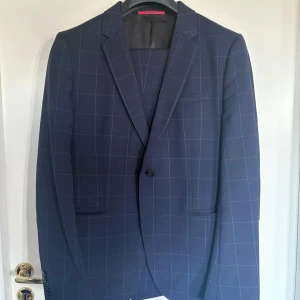 Mörkblå rutig kostymsett från Hugo - Säljer ett stilrent mörkblått kostymsett från Hugo med diskret rutmönster. Kavajen har klassisk krage och två fickor framtill. Perfekt för bal, arbete eller fest!