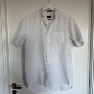 Vit linneskjorta från Gant - Säljer en klassisk vit kortärmad linneskjorta från Gant i regular fit. Skjortan har bröstficka, knappar framtill och en stilren krage. Perfekt för varma dagar och enkel att matcha med olika outfits. Finns fläckar under ärmarna men går säkert att få bort med en tvätt. Övrigt bra skick