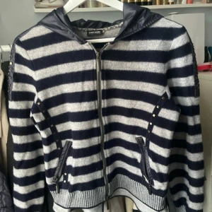 Randig hoodiejacka  - Mysig hoodiejacka med breda marinblå och grå ränder, dragkedja framtill och huva i glansigt material. Jackan har dekorativa nitar längs fickor och ärmar samt ribbade muddar. Jackan/koftan köpte ja för 2 tusen och den är i ull, luvan går att justera. Den säljs inte längre!
