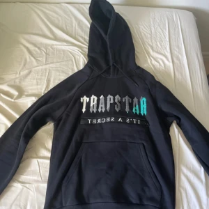 HEL TRACKSUIT TRAPSTARR  - Säljer en svart hoodie från Trapstar med stor broderad logga i vitt, grått och turkost på bröstet. Huvan har en turkos broderad detalj och tröjan har en magficka samt snörning i huvan. Perfekt för dig som gillar streetwear.