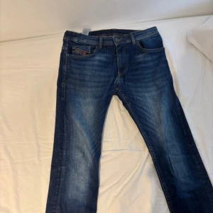 Blå jeansbyxor från Diesel - Säljer ett par klassiska blå jeans från Diesel med snygga slitningar och raka ben. Jeansen har fem fickor, gylf med dragkedja och knapp samt tydlig Diesel-logga på bakfickan och myntfickan. Perfekta för en avslappnad stil.
