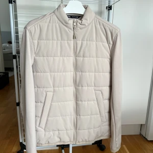 Beige quiltad cardiganjacka med ärmar, från Zara - Stilren beige quiltad cardiganjacka från Zara med dragkedja framtill och två snedställda fickor. Cardiganjacka har en hög krage och är tillverkad i ett mjukt syntetmaterial. Perfekt för lager-på-lager under kyligare dagar.