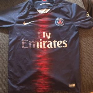 PSG hemmatröja Nike - Säljer en blå Paris Saint-Germain fotbollströja från Nike med röd och vit detalj på bröstet och klubbmärke på vänster sida. Tröjan har korta ärmar och trycket 'Fly Emirates' på framsidan. Perfekt för fans av PSG.