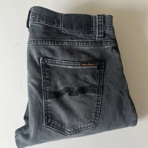 Svarta jeans från Nudie Jeans - Säljer ett par svarta jeans från Nudie Jeans med klassisk femficksdesign och broderad bakficka. Jeansen har en rak passform och är tillverkade i denim med lätt tvättad look.