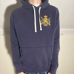 Mörkblå hoodie från Polo Ralph Lauren - Säljer en mörkblå hoodie från Polo Ralph Lauren med gul broderad logga på bröstet. Tröjan har vit dragsko i huvan och en stor magficka framtill. Perfekt för en avslappnad stil.