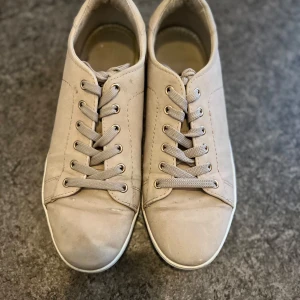 Beige sneakers med snörning - Säljer ett par stilrena beige sneakers med rund tå och platt vit sula. Skorna har klassisk snörning och är tillverkade i syntetmaterial med diskreta sömmar på sidorna. Perfekta för en clean och enkel look.