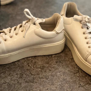 Vita sneakers med platåsula och dragkedja - Säljer ett par vita sneakers med platåsula och snörning. Skorna har även en dragkedja på sidan för enkel på- och avtagning. Klassisk rund tå och diskret design som passar till mycket. Perfekta för dig som gillar stilrena och bekväma skor.