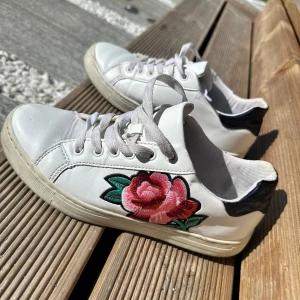 Vita sneakers med broderad ros - Säljer ett par vita sneakers med broderad rosa och röd ros på sidan. Skorna har svarta detaljer vid hälen och vita snören. Perfekta för dig som vill ha en unik och färgglad detalj till outfiten.