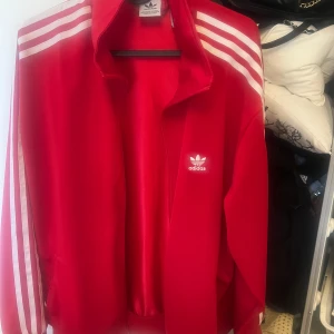 Röd Adidas track jacket med vita ränder - Säljer en klassisk röd Adidas track jacket med vita ränder längs ärmarna och Adidas-logga på bröstet. Jackan har dragkedja och hög krage, perfekt för en sportig och retro look.