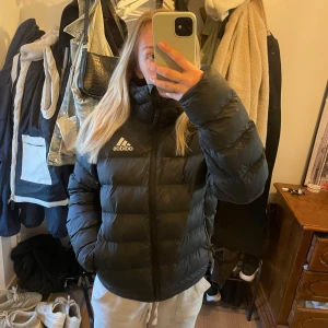 Svart pufferjacka från Adidas - Säljer en svart pufferjacka från Adidas med huva och dragkedja. Jackan har Adidas-logga på bröstet och är tillverkad i polyester. Perfekt för kyliga dagar och har en sportig look.