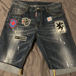 Jeansshorts med patches från Dsquared2 - Snygga jeansshorts från Dsquared2 med flera coola patches och slitningar. Modellen är slim och har uppvikta benslut. Mörkblå denim med detaljer som broderade märken och tryck både fram och bak.